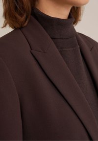 Zbliżenie na osobę noszącą ciemno-brązowy sweter z golfem pod dopasowan ą marynarką, ukazujące okolicę szyi i ramion.