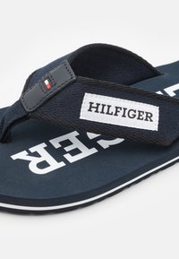 Marine flip-flop med en tekstureret stofrem, hvid logo-patch og en polstret sål med hvide accenter langs kanten.