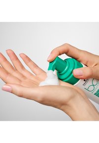 Un produit capillaire moussant est distribué d'un flacon-pompe vert dans une main. La mousse est blanche, et le flacon porte une étiquette blanche avec un texte vert.