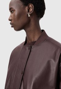 Hnědá kožená bombardérka s texturovanou přední částí a zapínáním na patentky. Hladký povrch, vroubkovaný límec a moderní design.