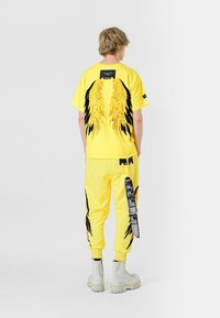 Chándal amarillo con gráficos de alas y acentos negros, que incluye una camiseta de manga corta y pantalones con puños, combinados con botas blancas.
