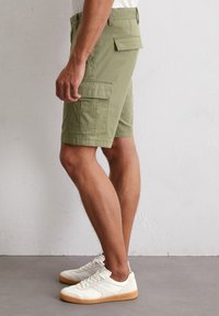 Olivgrüne Cargo-Shorts aus strukturiertem Material, mit mehreren Seitentaschen und einem einfachen Bunddesign. Getragen mit weißen Sneakers.