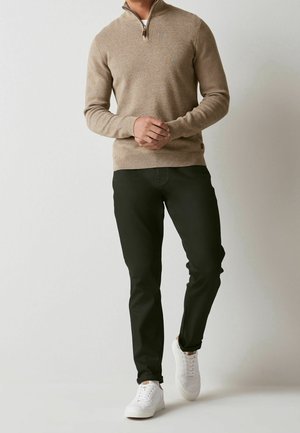 Beige zip-up sweater with a textured finish, paired with fitted black trousers and white sneakers. Sweater features a stand collar detail.