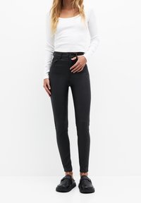Pantalon skinny taille haute noir en tissu lisse et extensible ; associé à un haut blanc cintré à manches longues et des chaussures noires sans lacets.
