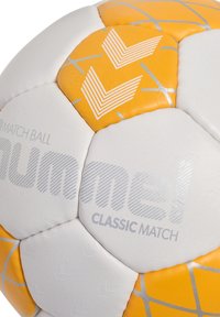 Biała i pomarańczowa piłka nożna z teksturowanymi panelami, geometrycznymi wzorami i wypukłym brandingiem "Classic Match" w srebrnym kolorze.