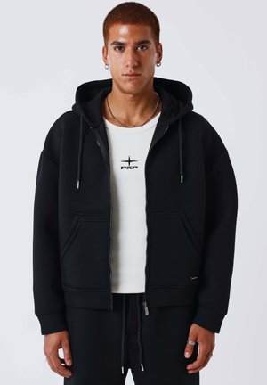 Jeune homme aux cheveux courts portant un sweat à capuche noir zippé sur un T-shirt blanc avec un logo étoile, debout devant un fond clair uni.