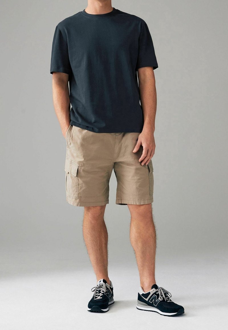 Homme portant un t-shirt noir, un short cargo beige et des baskets noires et blanches, debout devant un fond gris uni.