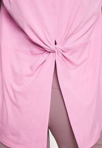 Chemise rose nouée dans le dos, révélant des leggings mauves en dessous.