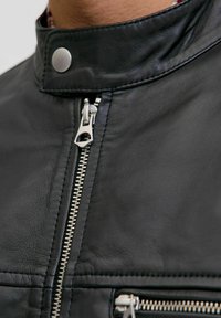 Detailní záběr na černou koženou bundu se zapínáním na zip a stříbrném zipu, včetně klopy s drukem.