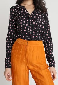 Blusa nera con piccolo motivo floreale rosa, caratterizzata da scollatura a V e chiusura con bottoni; abbinata a pantaloni arancioni a pieghe, design a vita alta.