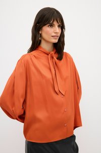 Rostorange Bluse mit gebundenem Kragen, langen Ärmeln und Knopfverschluss. Glattes Material mit leichtem Glanz und subtilen Schulterfalten.