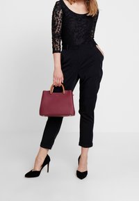 Schwarzes Spitzenoberteil mit langen Ärmeln, maßgeschneiderte dunkle Hose, schwarze Wildleder-Pumps und eine bordeauxrote Lederhandtasche mit hellbraunen Griffen.