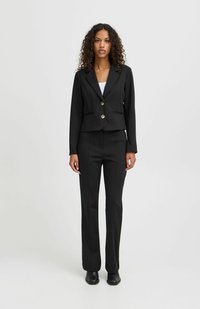 Schwarzer, maßgeschneiderter Anzug mit einem verkürzten Blazer und goldenen Knöpfen, Fronttaschen und schmal geschnittenen Hosen. Glattes Material mit taillierter Passform.