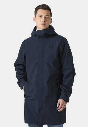 Joven con un largo impermeable azul marino con capucha, de pie con las manos en los bolsillos, fondo liso y claro.