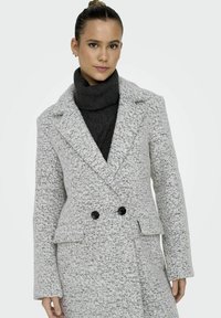 Manteau croisé en gris clair, tissu texturé avec des taches sombres. Présente des revers et des boutons noirs, associé à un col roulé sombre.