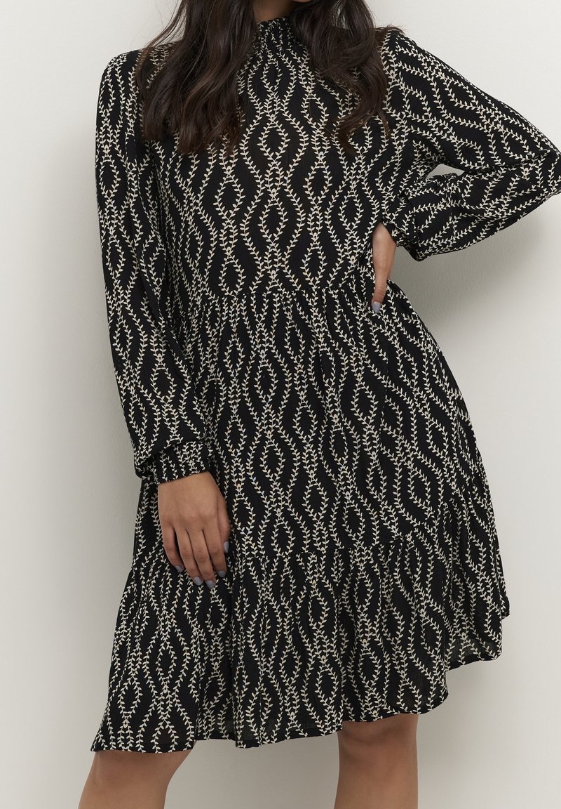 Vestido negro con manga larga, cuello alto y estampado de hojas blancas. La cintura fruncida y la falda de capas crean una silueta fluida.