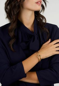 Bracciale dorato dal design fluido con una forma astratta. Indossato su un braccio nudo, in contrasto con una blusa blu navy.
