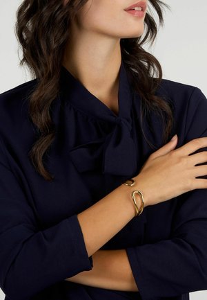 Bracciale dorato dal design fluido con una forma astratta. Indossato su un braccio nudo, in contrasto con una blusa blu navy.