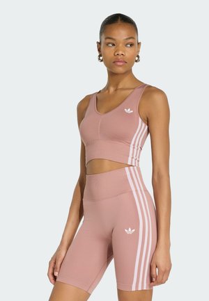 Donna che indossa un crop top sportivo mauve coordinato e pantaloncini da ciclismo a vita alta con strisce bianche e logo Adidas, in piedi davanti a uno sfondo semplice.