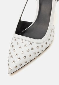Scarpa bianca con tacco alto e punta affusolata, caratterizzata da grandi occhielli in argento sulla superficie esterna e un design con tacco intagliato. Texture liscia.