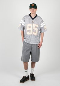 Joven con una camiseta deportiva gris con patrón y número 95, pantalones cortos grises con rayas finas, calcetines blancos, zapatos negros y gorra verde oscuro con logo naranja.