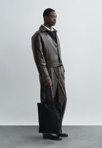 Rjava usnjena jakna z gumbom in žepom. Ujemajoče se hlače, črna torba z dolgimi ročaji in črne čevlje. Nevtralna ozadje.