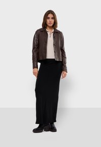 Veste en cuir marron avec un col et des boutons sur le devant, portée sur un pull crème. En dessous, une longue jupe noire et des bottes à talons noires.