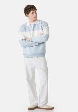 Mężczyzna stoi ze skrzyżowanymi ramionami, ubrany w jasnoniebiesko-biały sweter polo, białe spodnie i białe buty z brązowymi podeszwami.