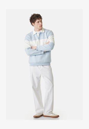 Man staat met de armen gekruist, draagt een lichtblauwe en witte polotrui, witte broek en witte schoenen met bruine zolen.