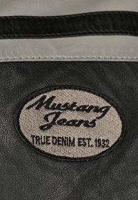 Svart läderlappar med broderad text "Mustang Jeans" i kursiv ovanför "True Denim Est. 1932" på en beige bakgrund.