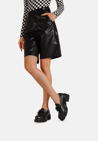 Pantaloni pantaloncini neri in ecopelle con vita alta, tasche e una texture liscia, abbinati a scarpe slip-on nere chunky con dettagli floreali.