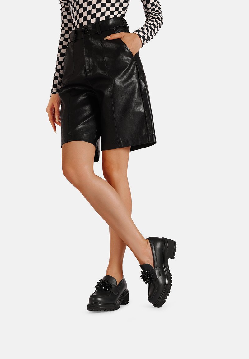 Pantaloni pantaloncini neri in ecopelle con vita alta, tasche e una texture liscia, abbinati a scarpe slip-on nere chunky con dettagli floreali.