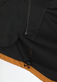 Giacca di tessuto nero con orlo inferiore arancione, caratterizzata da una cerniera, chiusura con coulisse e texture liscia. I dettagli includono accessori in metallo.