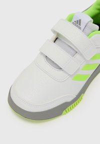 Zapatilla sintética blanca con dos tiras de Velcro texturizadas, suela gris y acentos verdes brillantes en el lado y el talón.
