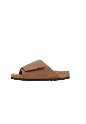 Sandalo slide in camoscio marrone con cinturino in velcro regolabile, plantare in sughero e suola in gomma nera, mostrato di profilo su sfondo bianco.