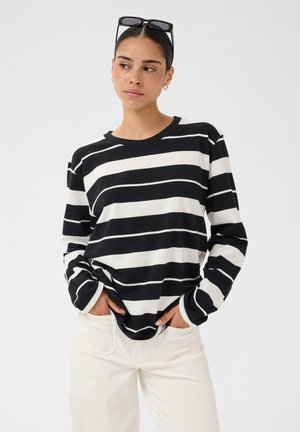 Femme aux cheveux foncés portant un t-shirt à manches longues rayé horizontalement noir et blanc, un pantalon blanc et des lunettes de soleil posées sur sa tête.