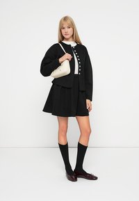 Svart kort cardigan med hvite knapper, båret over et svart skjørt. Modellen holder en liten beige veske, paret med høye svarte sokker og mørke sko.