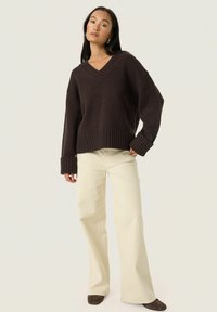 Pull brun oversized à col en V avec des poignets côtelés, associé à un pantalon large crème. Sa texture douce et sa coupe décontractée créent un look casual.