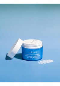Blauwe pot van Lumene Intense Hydration Moisturizer met een witte deksel, gedeeltelijk open, bevat een witte crème, naast een lepel crème op een blauwe achtergrond.