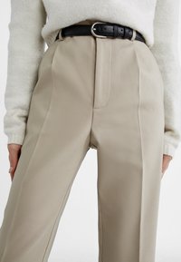 Beige op maat gemaakte broeken met een hoge taille, voorplooien en een zwarte leren riem met een zilveren gesp. Gladde stofstructuur is zichtbaar.