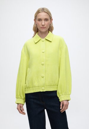 Γυναίκα με ανοιχτά ξανθά μαλλιά φοράει μια φωτεινή κίτρινη υφασμάτινη ζακέτα με μεγάλα κουμπιά και σκούρο τζιν μπροστά σε απλό λευκό φόντο.