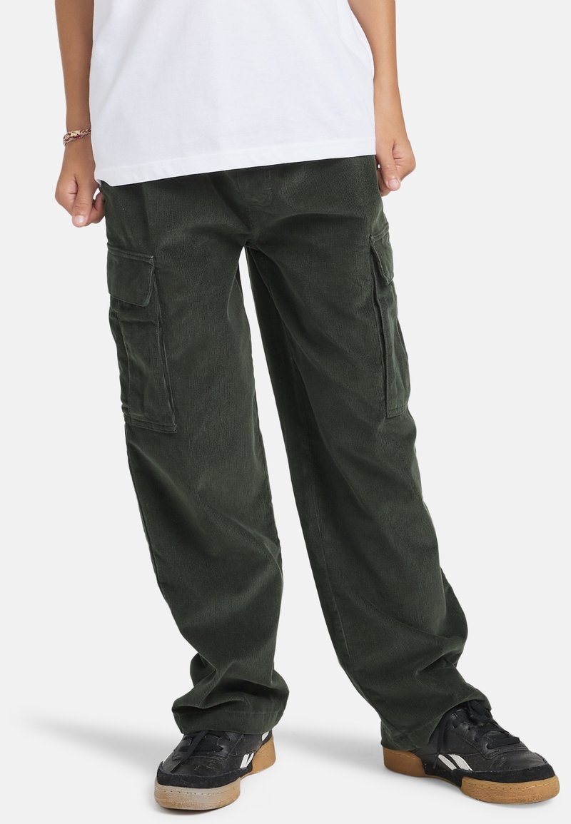 Personne portant un pantalon cargo vert foncé avec des poches latérales, des baskets noires et marron, et une chemise blanche, debout les mains détendues le long du corps.