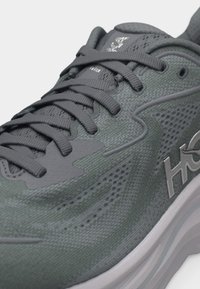 Κοντινό πλάνο γκρι-πράσινου παπουτσιού τρεξίματος με υφή δίχτυ επάνω μέρος, παχιά σόλα και γκρι κορδόνια δεμένα σε φιόγκο.