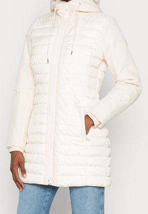 Kobieta w długiej kremowej pikowanej kurtce z kapturem, zamkiem z przodu, kieszeniami po bokach i jasnoniebieskich jeansach.