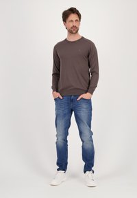 Felpa marrone a maniche lunghe con polsini a coste, abbinata a jeans chiari strappati e scarpe da ginnastica bianche. Il modello sta in piedi con le mani nelle tasche.