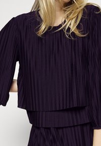 Lila plisserad topp med korta ärmar, lagerdesign och en mjuk textur. Har vertikala linjer och en löst sittande silhuett.
