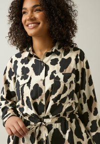 Die Button-Down-Bluse hat ein schwarzes und beigefarbenes Herzmuster, lange Ärmel, zwei Brusttaschen und ein gebundenes Taillendetail. Aus weichem, strukturiertem Stoff.