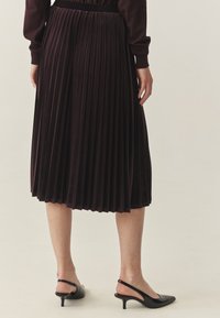 Donker bordeauxrode plooienrok van zachte stof, met een lengte tot halverwege de kuit en een elastische tailleband. Draagd met zwarte gespen met hoge hakken.