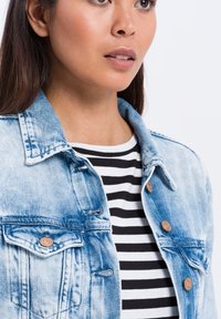 Jasnoniebieska kurtka dżinsowa z klasycznym kołnierzem, dwoma przednimi kieszeniami i miedzianymi guzikami, noszona na czarno-białą bluzkę w paski.