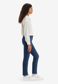 Žena s dugom kovrčavom kosom, odjevena u bijelu prugastu bluzu, plave traperice i bijele tenisice, stoji u profilu na bijeloj pozadini.